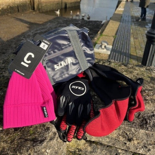 Cold weather gear including a pink beanie, gray gloves, black gloves, and red mittens on a stone surface with a harbor background.